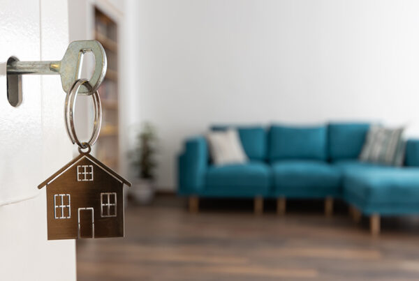 Steps for Buying Your Home - Close Up of Key with House Shaped Key Chain in the Door with a Couch and Living Space in the Background